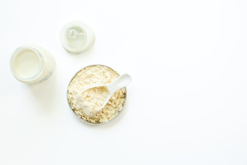 Powdered infant formula in jar lid and scoop with baby milk bottle on white background. Top view.