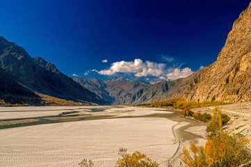 View of the wide, sandy riverbed contrasting with rugged, mountainous terrain under a deep blue sky, golden foliage adding warmth, Khaplu, Gilgit Baltistan, Pakistan.