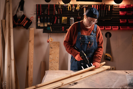 Female carpenter in overalls using cordless drill screwdriver in wooden furniture workshop - Powered by Adobe