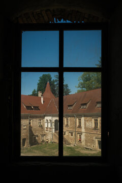 View of the old Bethlen Castle, framed by a dark window, reveals weathered stone walls and red tile roofs under a clear blue sky, Cris, Transilvania, Romania.