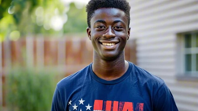 Smiling young man in a blue shirt with a flag design stands outdoors, showcasing a cheerful expression as he poses for the camera in a bright, natural setting