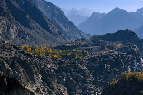 View of rugged, rocky terrain gives way to a small patch of golden autumn trees nestled amidst towering mountains under a clear blue sky, Jaglot, Gilgit Baltistan, Pakistan.