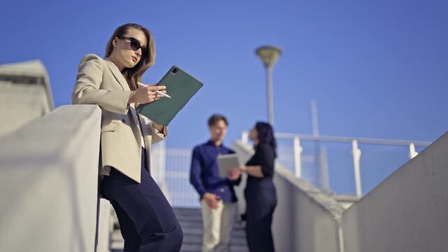 Confident businesswoman uses a digital tablet outdoors, her team collaborating behind her. Modern business people leverage technology for productivity and success under a clear blue sky.
