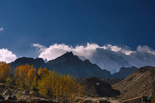 View of a dramatic landscape where rugged mountains rise, some capped with snow, against a deep blue sky dotted with clouds, and golden trees in Hunza Nagar, Gilgit Baltistan, Pakistan.