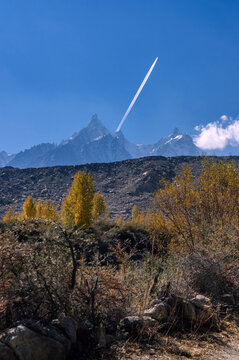 View of a jet stream piercing the deep blue sky above the snow-capped mountains, with golden trees dotting the rugged terrain, Gulmit, Gilgit Baltistan, Pakistan.