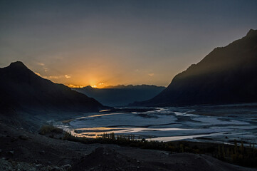 View of the sun breaking over the distant mountain peaks, illuminating the braided river below with a golden hue, contrasting against the dark silhouettes, Kachora, Gilgit Baltistan, Pakistan.