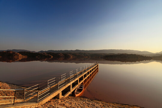 View of a long pier stretching into a tranquil lake reflecting the warm hues of the setting sun, with hills in the distance, Chakwal, Punjab, Pakistan.