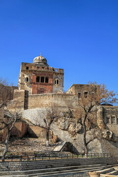 View of ancient architecture with weathered stone walls, a domed structure, and bare trees under a clear blue sky, Katas, Punjab, Pakistan.