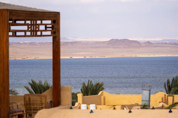 View of calm blue waters meeting the arid desert horizon under a muted sky, framed by the warm tones of local architecture, Qaroun Protected Area, Faiyum Governorate, Egypt.