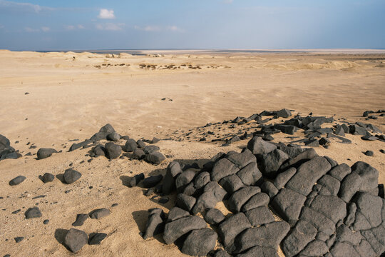 View of dark, angular rocks scattered across the sun-baked, sandy foreground, leading to a hazy horizon in the desert landscape, Qaroun Protected Area, Faiyum Governorate, Egypt.