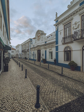 View of the cobblestone street leading to the vintage white buildings with blue trim under a cloudy sky, Ericeira, Lisbon, Portugal.