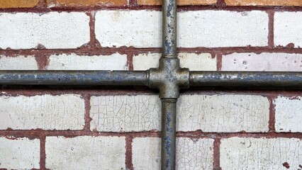 A close-up architectural detail of weathered grey metal pipes forming a cross joint against an old, cracked cream brick wall with reddish-brown grout lines and aged texture.