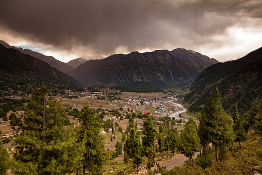 View of a valley town nestled between towering mountains under a dramatic sky, where lush green trees meet rugged peaks, Kalam, Khyber Pakhtunkhwa, Pakistan.