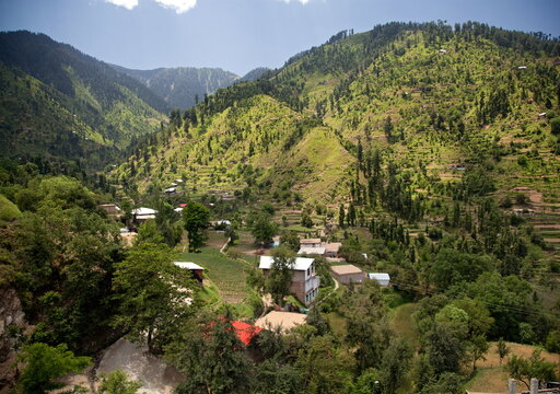 View of verdant hillsides embrace scattered dwellings under a bright sky, a serene landscape of natural beauty and human settlement, Kalam, Khyber Pakhtunkhwa, Pakistan.