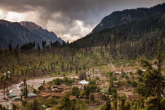 View of a village nestled between lush green hills and a flowing river under a dramatic sky, a serene landscape in Kalam, Khyber Pakhtunkhwa, Pakistan.