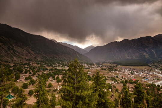 View of a valley town nestled among towering mountains under a dramatic, stormy sky, the landscape alive with vibrant greens and earthy tones, Kalam, Khyber Pakhtunkhwa, Pakistan.