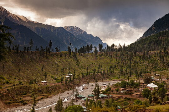 View of a river winding through a valley, flanked by verdant hills dotted with trees and small buildings, beneath a dramatic sky, Kalam, Khyber Pakhtunkhwa, Pakistan.