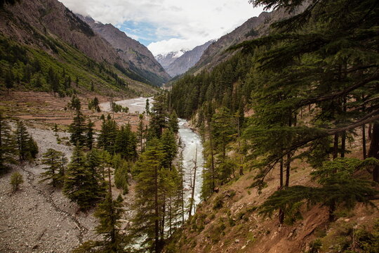 View of a winding river cutting through a verdant valley, flanked by towering, rugged mountains under a cloudy sky, Kalam, Khyber Pakhtunkhwa, Pakistan.