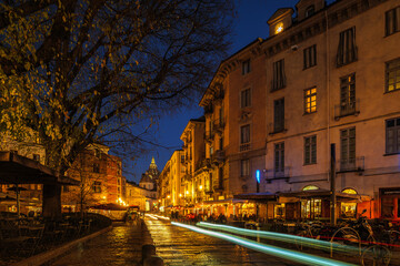 Turin, Italy. Night view of Turin Duomo from Largo IV Marzo, with glowing street lights, outdoor cafes, people and car light trails, capturing historic architecture and urban nightlife.