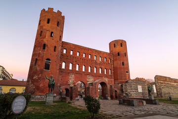 Turin, Italy. Porta Palatina, ancient Roman gate of Augusta Taurinorum in Turin at sunset, with red brick towers, arches and statues standing under a clear pastel sky.