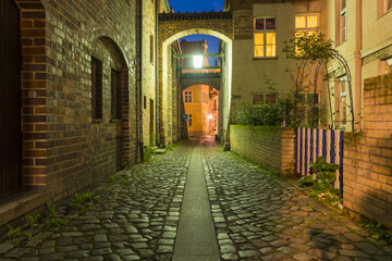 View of cobblestone street weaving through historic brick buildings under archways glowing with warm light, Stralsund, Mecklenburg-Vorpommern, Germany.