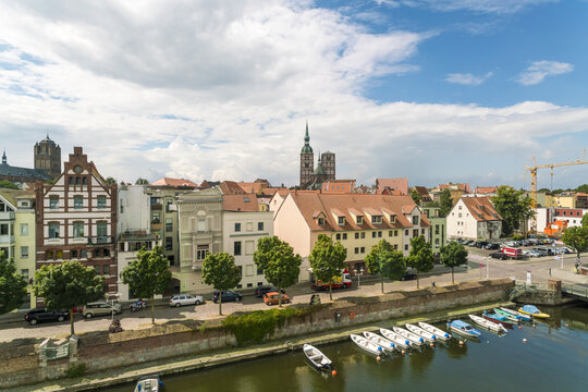 View of aged buildings with red-tiled roofs meet the serene waters, as bright boats bob gently, under a sky brushed with white clouds, Stralsund, Mecklenburg-Vorpommern, Germany.