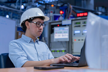 Asian male engineer wearing hardhat coding on laptop in industrial office. Professional technician...