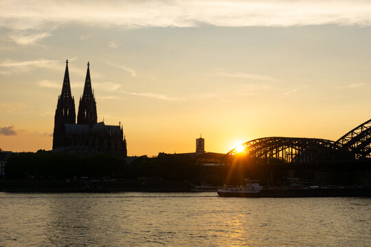 View of the sun setting behind the Hohenzollern Bridge and cathedral, casting a golden glow over the tranquil river, creating a striking contrast, Cologne, Nordrhein-Westfalen, Germany.