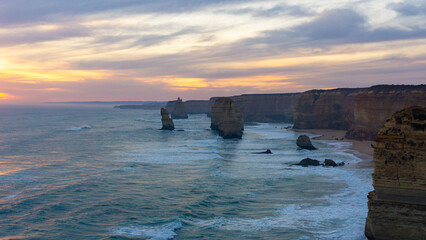 View of the rugged coastline where towering rock formations meet the turbulent sea under a sky painted with sunset hues, Twelve Apostles, Victoria, Australia.