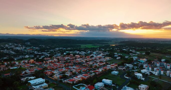 Flight over the red-roofed low-rise houses in the scenery of Ponce, Puerto Rico. Long cloud covering the setting sun at backdrop.