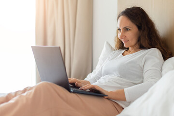 Adult woman reclining on bed using laptop in bright bedroom. Relaxed remote work concept, lifestyle balance, home office routine, modern technology, and comfortable interior
