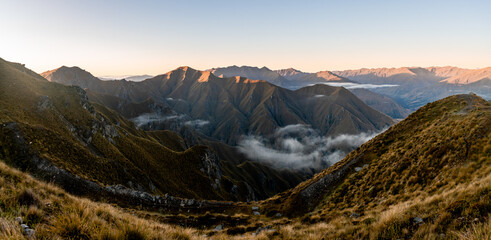 View of sunlit mountain peaks casting long shadows over valleys filled with ethereal clouds, creating a dramatic landscape, Roys Peak, Otago Region, New Zealand.