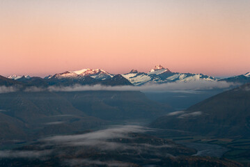 View of the snow-capped peaks kissed by the warm glow of the setting sun, hovering over valleys shrouded in ethereal mist, Roys Peak, Otago Region, New Zealand.