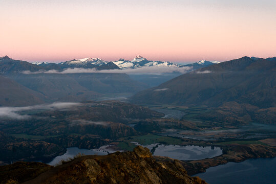 View of the rugged, sun-kissed foreground leading to a valley with a still lake, surrounded by majestic, snow-capped mountains under a soft, pastel sky, Roys Peak, Otago Region, New Zealand.