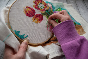 Woman’s hands embroidering colorful flowers using cross-stitch technique on fabric. Creative...