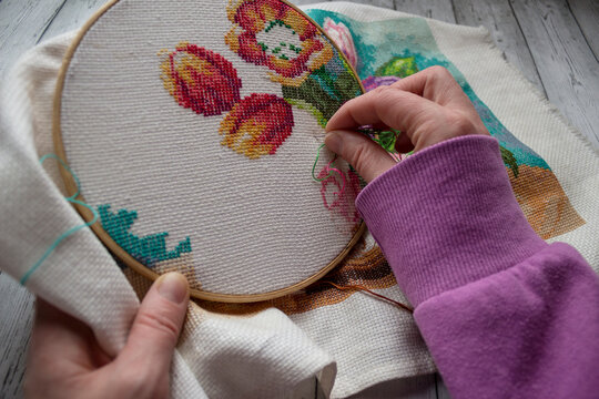 Woman&rsquo;s hands embroidering colorful flowers using cross-stitch technique on fabric. Creative handmade needlework process, perfect for DIY, craft hobby, textile art, creativity and slow handmade lifest