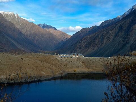 View of a placid, dark lake mirroring the majestic, snow-dusted mountains and earthy tones of the landscape under a bright sky, Gupis, Gilgit Baltistan, Pakistan.