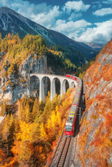 Aerial view of modern red train on Landwasser viaduct in alpine mountains, colorful forest at sunset in autumn. Bernina Express, Switzerland. Top view of train, railroad, orange and red trees in fall