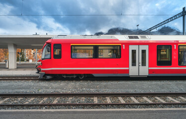 Red modern passenger train on mountain railway station in Swiss Alps in autumn. High-speed train, railway platform and cloudy sky. Bernina Express. Rail transport. Station in St. Moritz, Switzerland