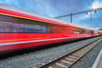 Blurred red passenger train passing mountain railway station in Swiss Alps at dusk. Moving high-speed train. St. Moritz, Switzerland. Bernina Express. Railway platform and cloudy sky. Rail transport