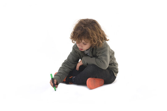 Side view of a child  sitting on the ground painting, drawing and looking down with a marker on a white background.