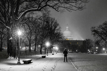 View of a silhouette stands on a snow-covered path beneath glowing streetlights, with the US Capitol dome softly illuminated in the distance, Washington DC, United States.