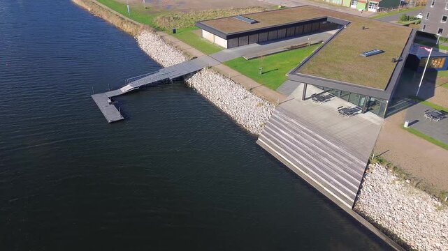 Modern waterfront architecture by fjord in southern Denmark. The scene shows contemporary public buildings with green roofs, stone steps, and a small pier meeting calm blue water