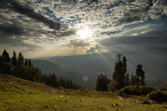 View of sunbeams pierce through dramatic clouds over rolling mountains and lush green slopes dotted with tall trees, Sri Paye, Khyber Pakhtunkhwa, Pakistan.