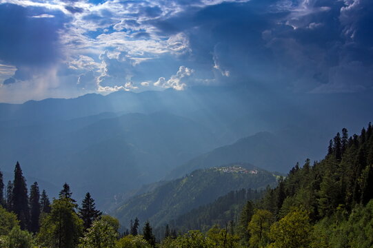 View of sun rays piercing through dramatic clouds over the misty mountains, casting a serene glow on the lush green landscape, Sri Paye, Khyber Pakhtunkhwa, Pakistan.