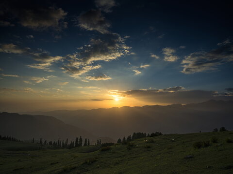 View of the sun setting behind hazy mountains casting long shadows over green hills and scattered trees, Sri Paye, Khyber Pakhtunkhwa, Pakistan.