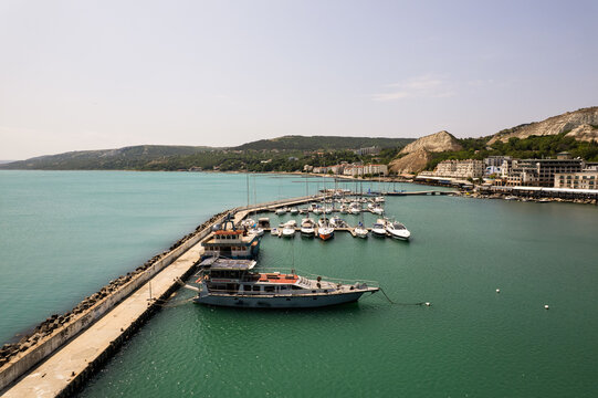 Aerial view of boats docked at the marina, where the turquoise water meets the concrete pier, framed by distant cliffs, Balchik, Dobrich Province, Bulgaria.