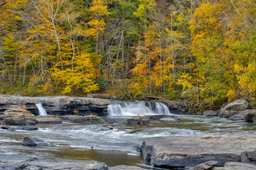  A tranquil waterfall flows gracefully over layered rock formations into a calm pool, framed by smooth stones and surrounded by a forest glowing with vibrant autumn foliage 