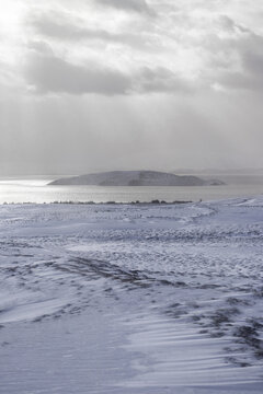 View of a snow-covered landscape leading to a distant island under a cloudy sky, the cold hues painting a serene yet stark scene, Iceland.