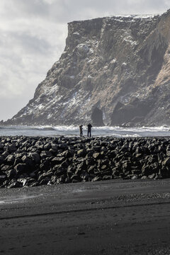 View of two figures standing on a dark rocky barrier against the backdrop of a massive cliff face dusted with snow, contrasting with the black sand beach, Vik, Iceland.